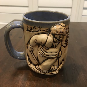 Puede incluir: Una taza de cerámica azul con un diseño en relieve marrón y blanco de una persona esquiando en una escena de invierno. La taza tiene un asa y un borde ancho.