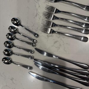 May include: A collection of stainless steel silverware, including forks, spoons, and knives. The utensils are arranged on a light-colored surface, showcasing their polished, reflective surfaces. The forks have four tines, and the spoons have a rounded bowl.