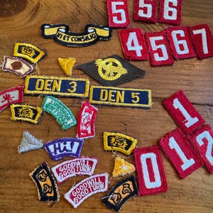 May include: A collection of vintage patches and badges in various shapes and colors. Includes patches with numbers, text, and graphic designs. The patches are in red, yellow, black, and white.