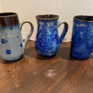 May include: Three ceramic mugs with unique designs. The mug on the left is tall with a brown top and blue accents. The middle mug has a blue floral pattern. The mug on the right is blue with a similar pattern. All have handles.