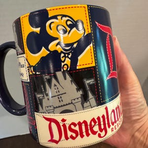 May include: A navy blue Disneyland Resort coffee mug featuring Mickey Mouse, a castle, and the Disneyland logo. The mug has a stitched design with yellow, red, and white accents.
