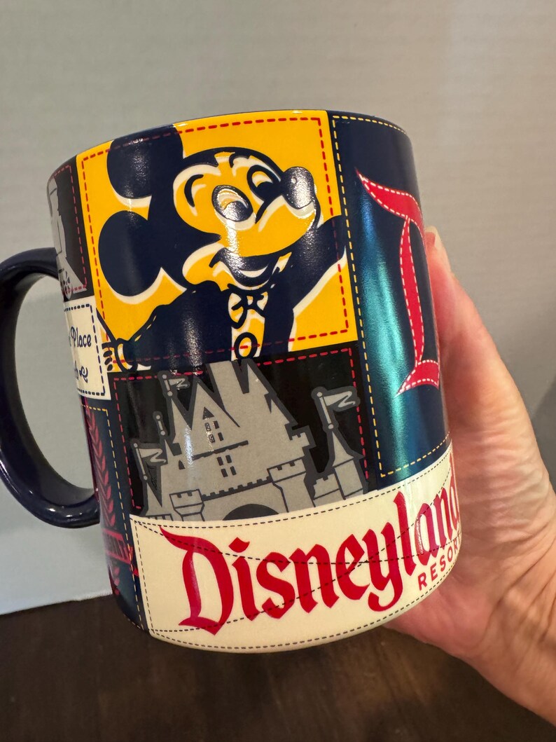 May include: A navy blue Disneyland Resort coffee mug featuring Mickey Mouse, a castle, and the Disneyland logo. The mug has a stitched design with yellow, red, and white accents.