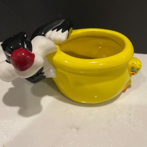 May include: A yellow ceramic planter shaped like Tweety Bird with Sylvester the Cat peeking out from the side. The planter is decorated with black and white details.