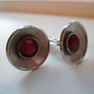 May include: A pair of silver cufflinks with red gemstone accents. The cufflinks have a circular design with a raised center and a textured outer edge.