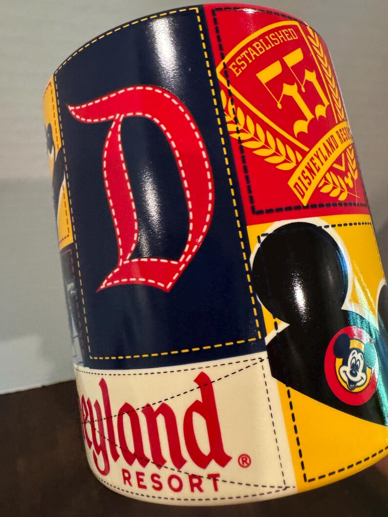 May include: A cylindrical souvenir cup featuring a patchwork design with the Disneyland Resort logo in red, a large red "D", and a Mickey Mouse silhouette. The cup has a yellow, blue, and red color scheme.