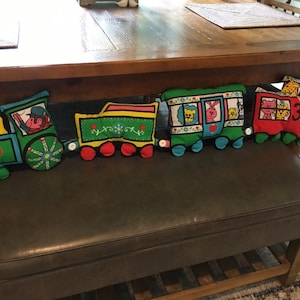 May include: A colorful train-themed pillow set with a red, yellow, and green engine, a green boxcar, a green car with a lion, a blue car with a rabbit, and a red car with a giraffe. The pillows are arranged in a line on a brown leather bench.