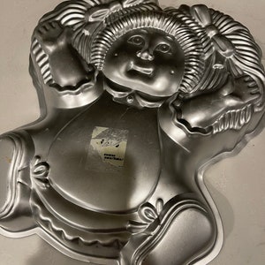 May include: A silver metal cake pan shaped like a doll with pigtails and a bow. The doll is wearing a dress and shoes.