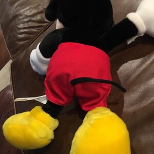May include: A plush Mickey Mouse toy with black fur, red shorts, and yellow shoes. The toy is lying on its back with its arms outstretched.