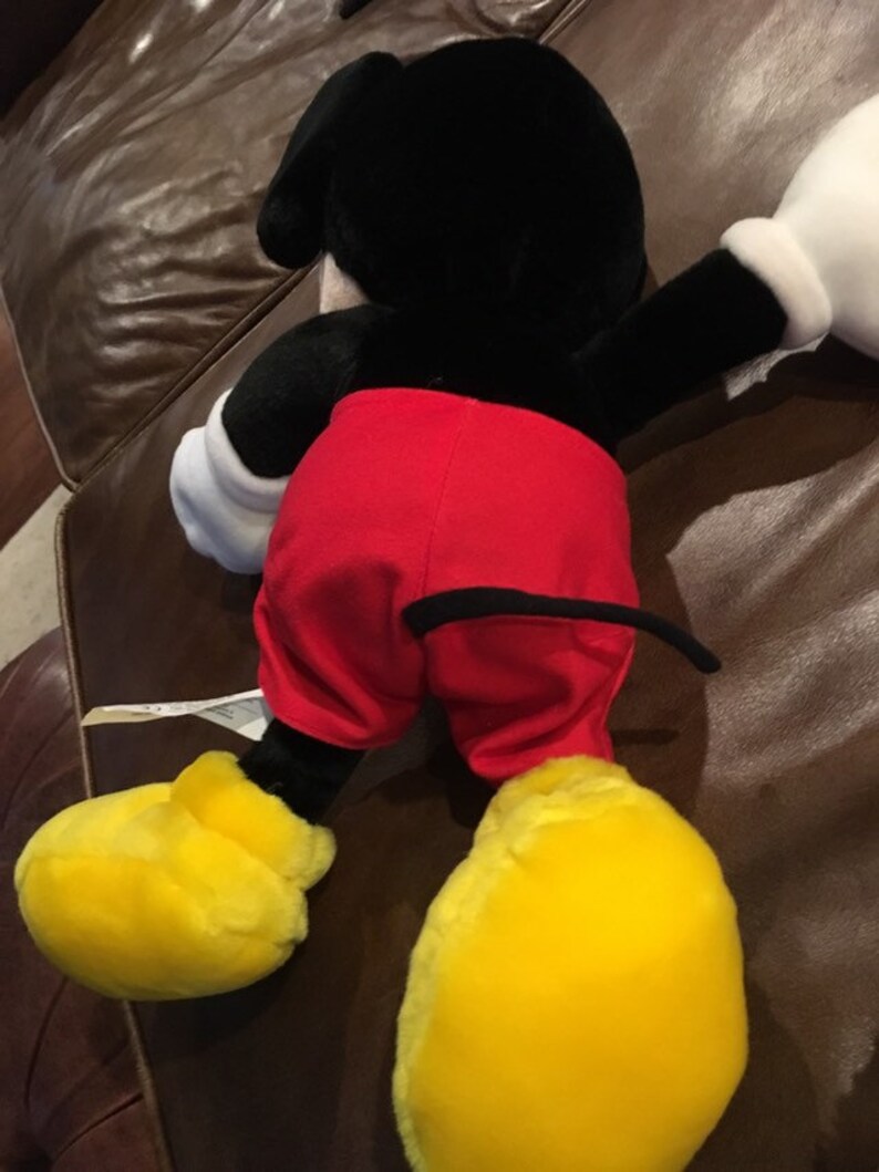 May include: A plush Mickey Mouse toy with black fur, red shorts, and yellow shoes. The toy is lying on its back with its arms outstretched.