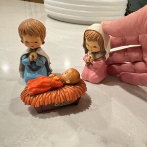 May include: A nativity scene figurine set with a baby Jesus in a manger, a girl with brown hair wearing a blue dress, and a girl with brown hair wearing a pink dress.