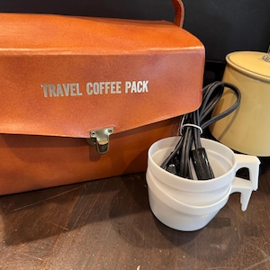 May include: A vintage travel coffee pack set. The set includes a brown leatherette case with "TRAVEL COFFEE PACK" printed on it, a yellow canister with a glass lid, a white cup, and a black power cord.