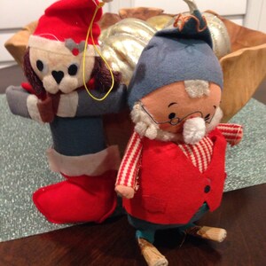 May include: Two vintage Christmas ornaments, one a red and white elf with a blue hat and white beard, the other a red and white dog wearing a Santa hat.