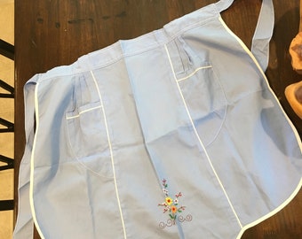 May include: A light blue apron with white trim and a scalloped hem. The apron has two pockets and a floral embroidery design.