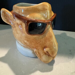 May include: A ceramic camel-shaped mug with brown sunglasses. The mug is tan with brown accents and a white base. The camel's head is tilted slightly, giving it a relaxed appearance. The sunglasses are dark brown with black lenses.