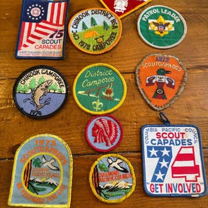 May include: A collection of vintage Boy Scout patches in various shapes and colors. The patches feature text like "Camp Cooper High Adventure", "Chinook District BSA Camporee", and "Scout Capades". Some patches have images of a fish, a Native American headdress, and a bird.
