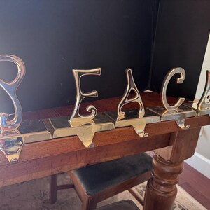 May include: A decorative metal stocking holder spelling out the word "PEACE". The letters are in a shiny gold color and sit on a rectangular base with hooks for hanging stockings. The holder is placed on a wooden table.