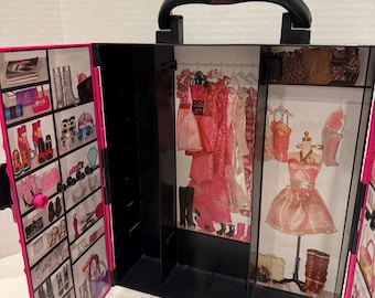 Puede incluir: Un armario Barbie negro con detalles en rosa. El armario se abre para revelar una variedad de ropa, zapatos y accesorios en miniatura. El asa es negra y el interior tiene una combinación de colores rosa y negro.