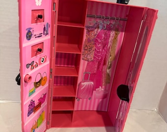 Puede incluir: Armario rosa para muñecas con asa negra y el nombre "Alisa". El armario está abierto, revelando estantes, una barra para colgar ropa con ropa de muñecas y una pequeña maleta. El lado izquierdo tiene estantes con accesorios en miniatura.