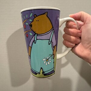 May include: A tall ceramic mug with a purple, blue, and green illustration of a bear wearing overalls and holding a flower. The bear is looking to the left and there is a small tree with red berries behind it.