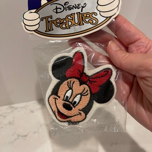 May include: A white fabric patch with an embroidered image of Minnie Mouse. Minnie Mouse is wearing a red bow and has a black nose and eyes. The patch is packaged in a clear plastic bag with a Disney Treasures header card.
