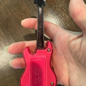 Puede incluir: Una peque&ntilde;a guitarra de juguete rosa con m&aacute;stil y clavijero negros. La guitarra tiene un compartimento para pilas en la parte trasera. La guitarra de juguete se sostiene en una mano. La guitarra es de color rosa brillante.
