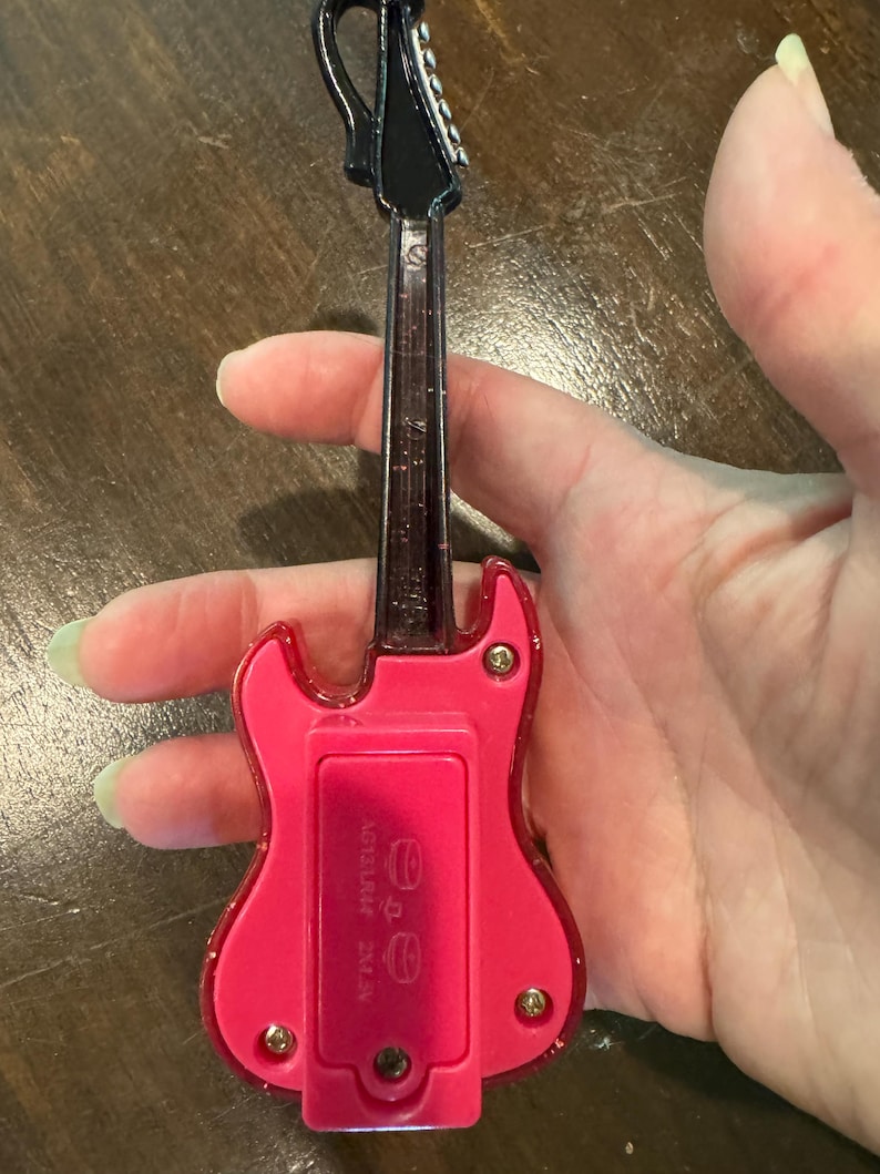 Puede incluir: Una peque&ntilde;a guitarra de juguete rosa con m&aacute;stil y clavijero negros. La guitarra tiene un compartimento para pilas en la parte trasera. La guitarra de juguete se sostiene en una mano. La guitarra es de color rosa brillante.