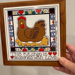 May include: A brown and white ceramic tile with a framed wooden border. The tile features a cartoon chicken with the text "This Kitchen is Seasoned with Love" and a copyright symbol with the year 1982 and the name Susan Marie McChesney.