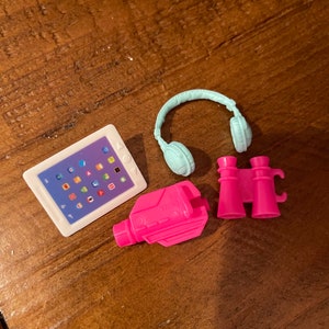 May include: A set of pink and blue toy accessories for a dollhouse or playset. The set includes a tablet, headphones, binoculars, and a camera.