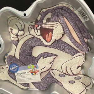 May include: A silver Bugs Bunny cake pan featuring a cartoon image of Bugs Bunny in a running pose. The pan is made by Wilton and has the text "Bugs Bunny" and "Fun ideas on our cake box" printed on it.