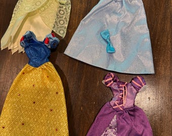 May include: Four doll dresses in various colors. One is yellow with lace, another is light blue with a bow, a third is yellow with red accents, and the last is purple with a floral pattern.