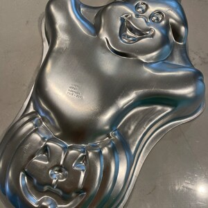 May include: A silver metal cake pan shaped like a ghost with a pumpkin on the bottom. The ghost has a smiling face and a hat. The text on the bottom reads "Made in Indonesia".