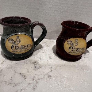 May include: Two ceramic mugs with handles, one dark blue and one dark brown. Each mug features an oval cream-colored decal with a black rooster illustration and the text "Over Easy Cafe, Sanibel Island, Florida" printed in black lettering.