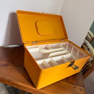 May include: An open, orange plastic toolbox with a white interior. The toolbox has a hinged lid and a metal clasp. The interior is divided into several compartments and slots for organization. The toolbox is sitting on a wooden table.