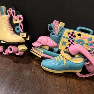 May include: Two vintage wall decor pieces in a retro 80s style. One features a yellow roller skate with pink wheels and a pink phone. The other features a blue sneaker with a yellow sole, a pink hat, and a yellow suitcase with a black and white checkered pattern.