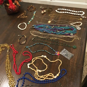 May include: A collection of necklaces and bracelets made of various materials, including beads, shells, and metal. The jewelry is arranged on a wooden table, with some pieces overlapping. The colors of the jewelry include blue, green, red, white, and gold.