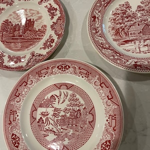 May include: Three red and white ceramic dinner plates with different patterns. The first plate has a red and white castle scene, the second plate has a red and white tree and house scene, and the third plate has a red and white willow tree pattern.