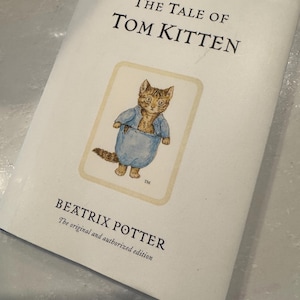 May include: The Tale of Tom Kitten book cover featuring a cartoon illustration of a kitten wearing a blue shirt and pants. The book is by Beatrix Potter, the original and authorized edition.