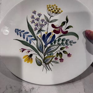 May include: White ceramic plate with a floral design. The design features a bouquet of flowers in shades of yellow, purple, blue, and burgundy, with green leaves. The plate has a simple, elegant design.