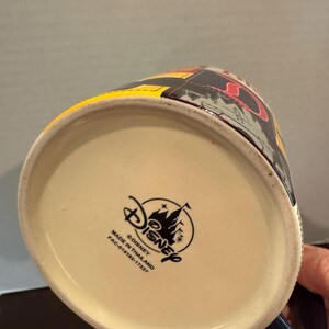 May include: A Disney ceramic mug with a colorful design. The mug has a cream-colored interior and a dark blue handle. The Disney logo and text are printed on the bottom of the mug. The mug is made in Thailand.