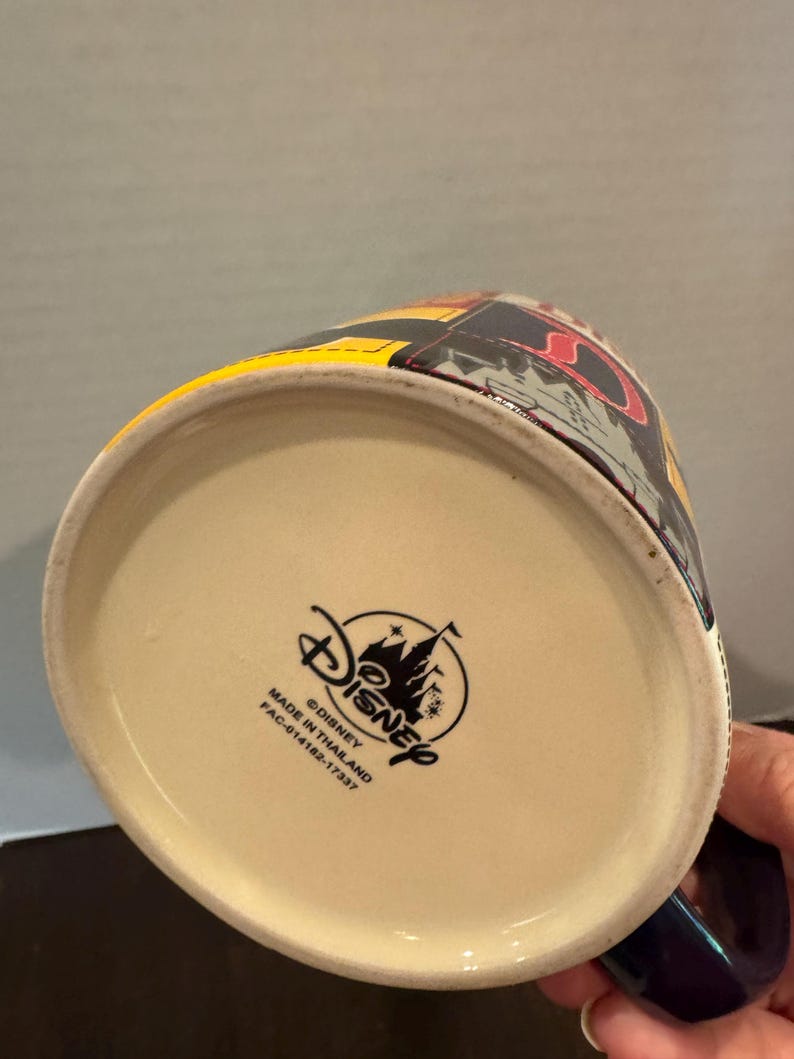 May include: A Disney ceramic mug with a colorful design. The mug has a cream-colored interior and a dark blue handle. The Disney logo and text are printed on the bottom of the mug. The mug is made in Thailand.