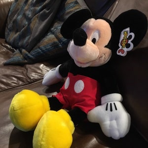 May include: A large plush Mickey Mouse toy sitting on a brown leather sofa. Mickey is wearing his signature red shorts with white polka dots and yellow shoes. He has a black nose and big black ears. The toy has a Mouseketeers 6 tag attached to his ear.