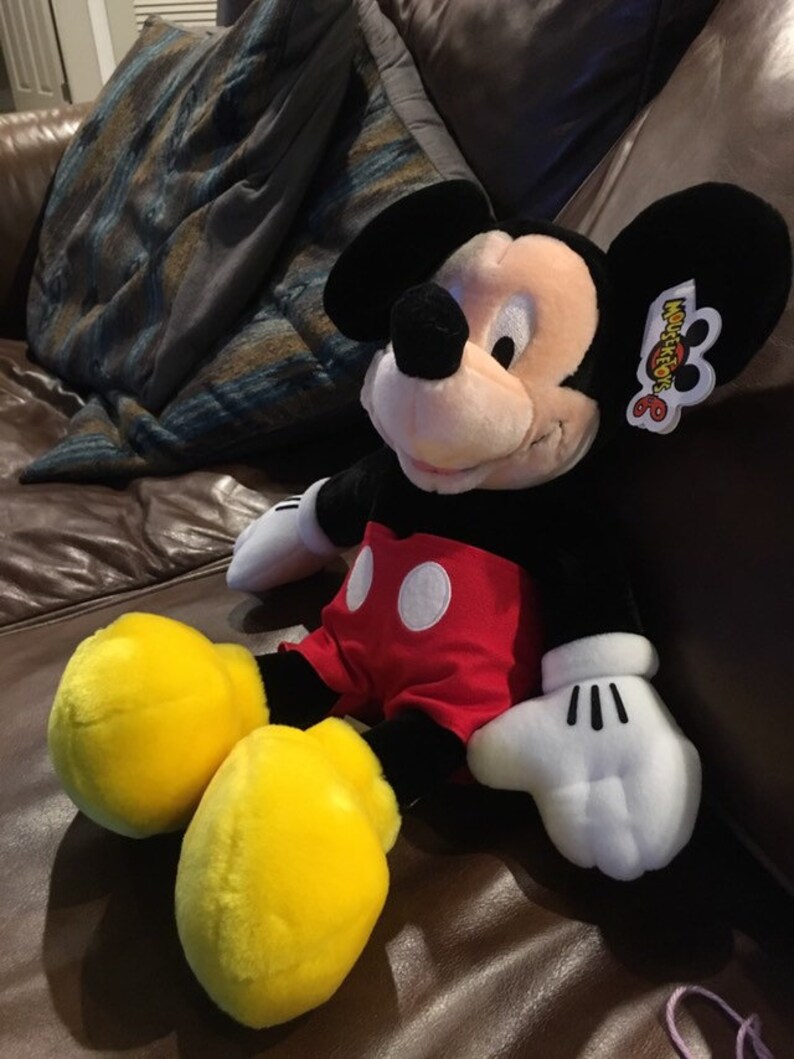 May include: A large plush Mickey Mouse toy sitting on a brown leather sofa. Mickey is wearing his signature red shorts with white polka dots and yellow shoes. He has a black nose and big black ears. The toy has a Mouseketeers 6 tag attached to his ear.