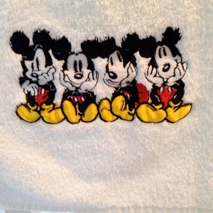 Puede incluir: Toalla de ba&ntilde;o blanca con un dise&ntilde;o bordado de cuatro personajes de Mickey Mouse en fila. Los personajes son negros con pantalones cortos rojos y zapatos amarillos.