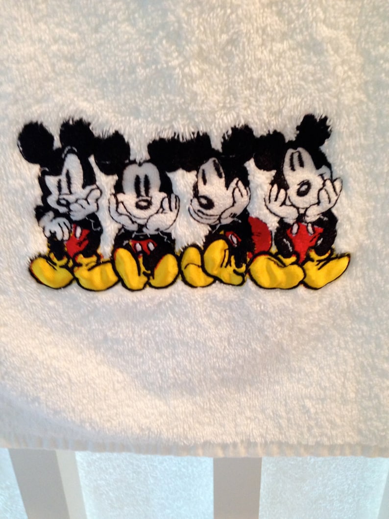 Puede incluir: Toalla de ba&ntilde;o blanca con un dise&ntilde;o bordado de cuatro personajes de Mickey Mouse en fila. Los personajes son negros con pantalones cortos rojos y zapatos amarillos.