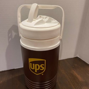 May include: A brown and white insulated beverage jug with a white lid and two handles. The jug has the UPS logo in gold on the front.