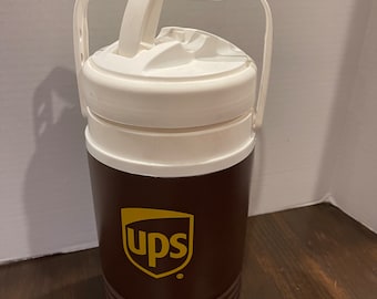 Op de afbeelding: Een bruine en witte geïsoleerde drankenkan met een wit deksel en twee handvatten. De kan heeft het UPS-logo in goud aan de voorkant.
