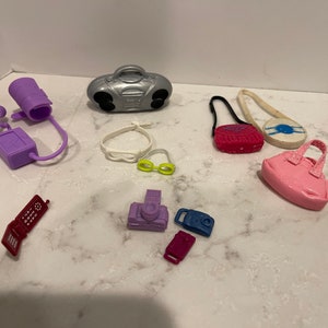 May include: A collection of miniature plastic accessories for a dollhouse, including a purple phone, a red phone, a purple and silver boombox, a purple purse, a pink purse, a white purse, a pair of yellow sunglasses, and a purple camera.