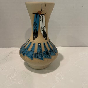 May include: A small, cream-colored ceramic vase with a blue and white feather design. The vase has a narrow neck and a wide base.