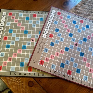 May include: Two brown and beige Scrabble game boards with the word "SCRABBLE" printed in red and white letters. The boards have a grid of squares with different colored squares for bonus points. The boards are on a wooden surface.