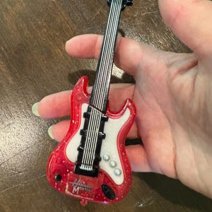 Puede incluir: Una guitarra roja y blanca en miniatura con el texto "Hannah Montana". La guitarra tiene cuerdas negras y un clavijero negro. El cuerpo de la guitarra es rojo con purpurina y un centro blanco.